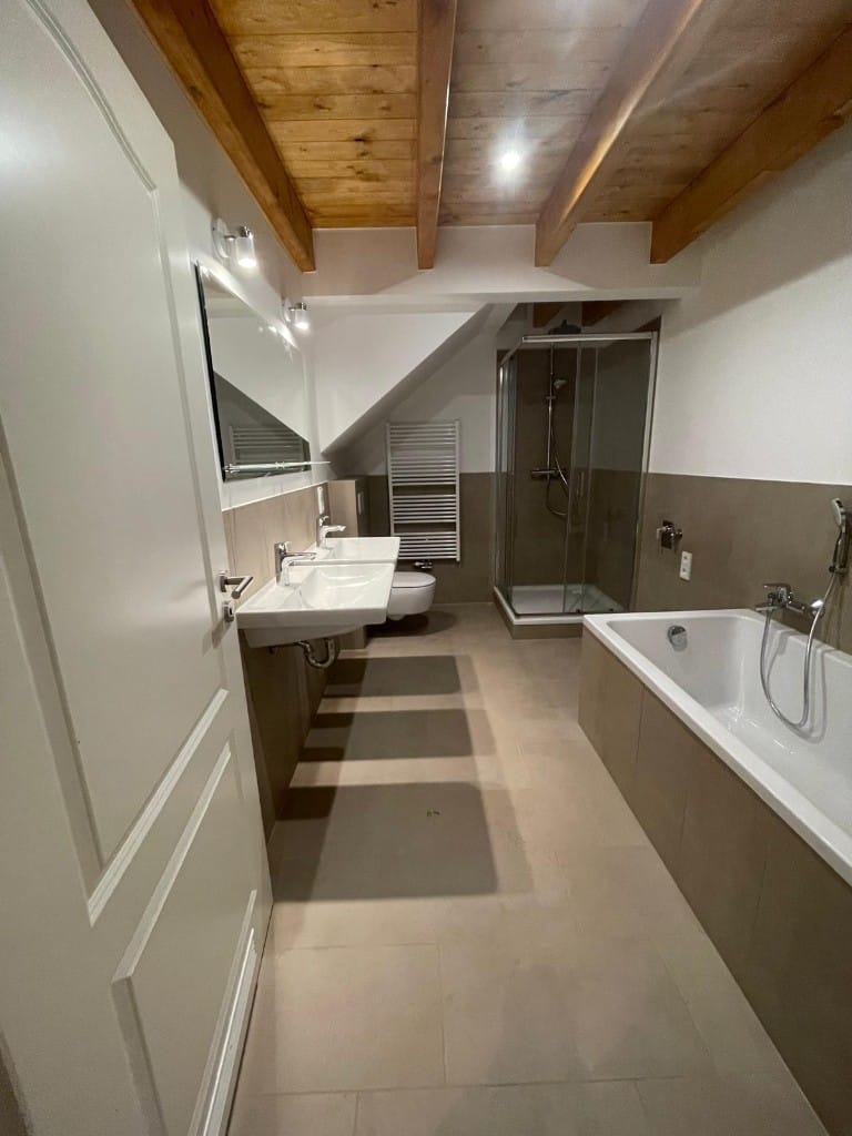 Blick ins fertige Bad: Doppelwaschtisch, Badewanne, Glasdusche und Holzbalkendecke.